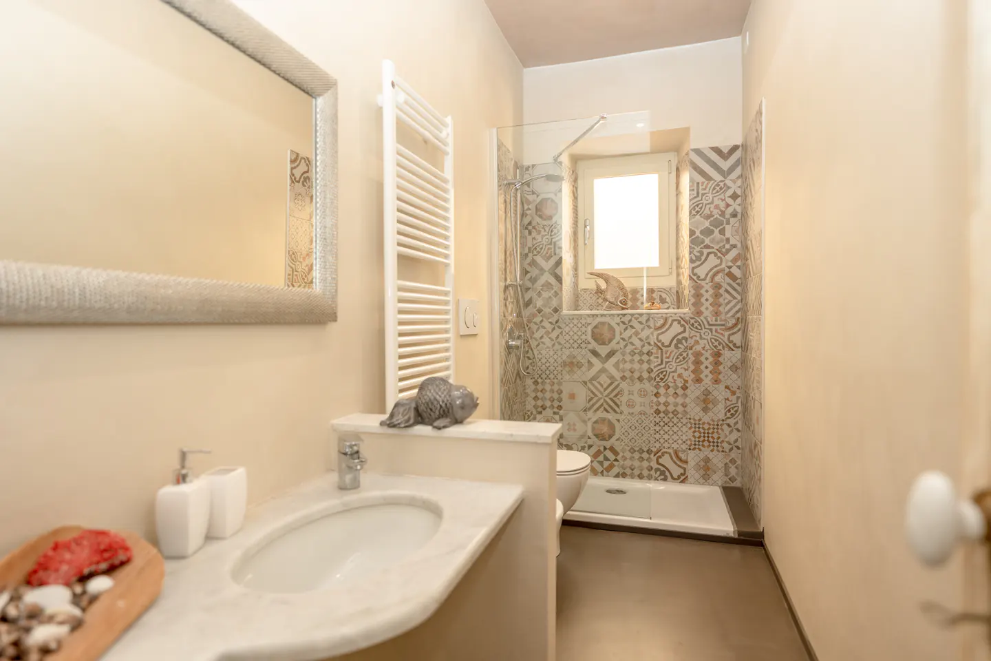 Bathroom with marble sink, silver mirror, and white towel rack. Shower with patterned tile and window in the background.