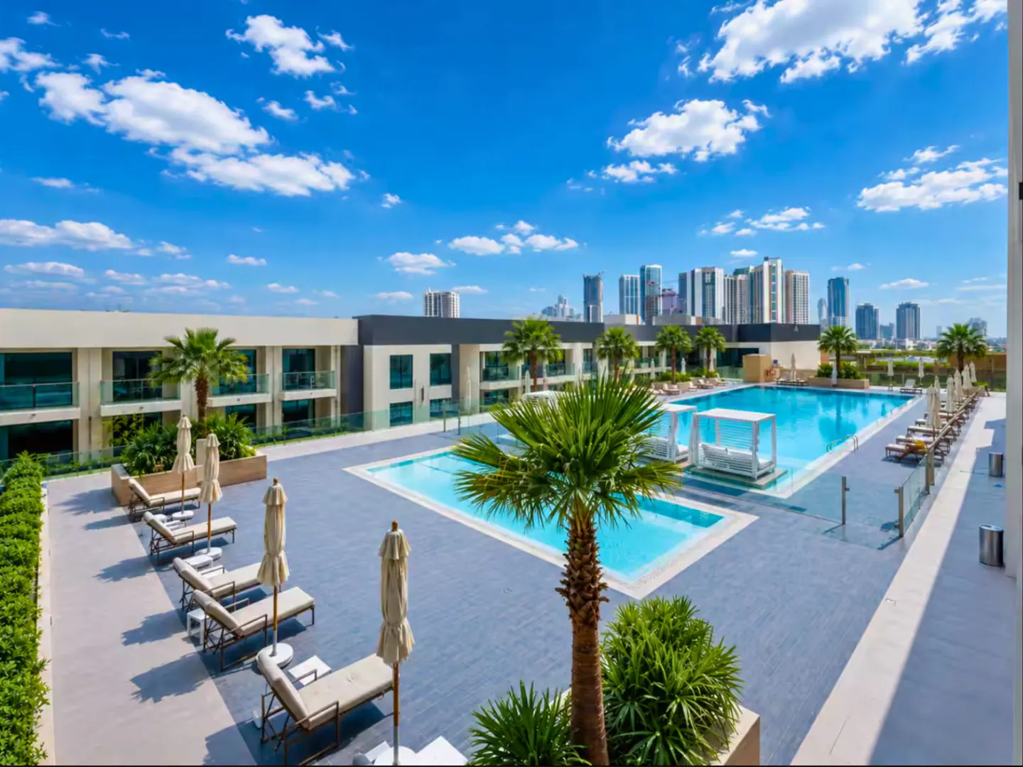 Rooftop pool area with lounge chairs, umbrellas, and palm trees. City skyline in the background under a blue sky with white clouds.