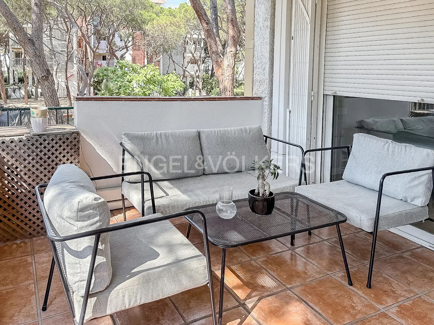 Balcony with gray cushioned sofa, chairs, and metal table. A small plant and glass vase sit on the table. Trees are visible in the background.
