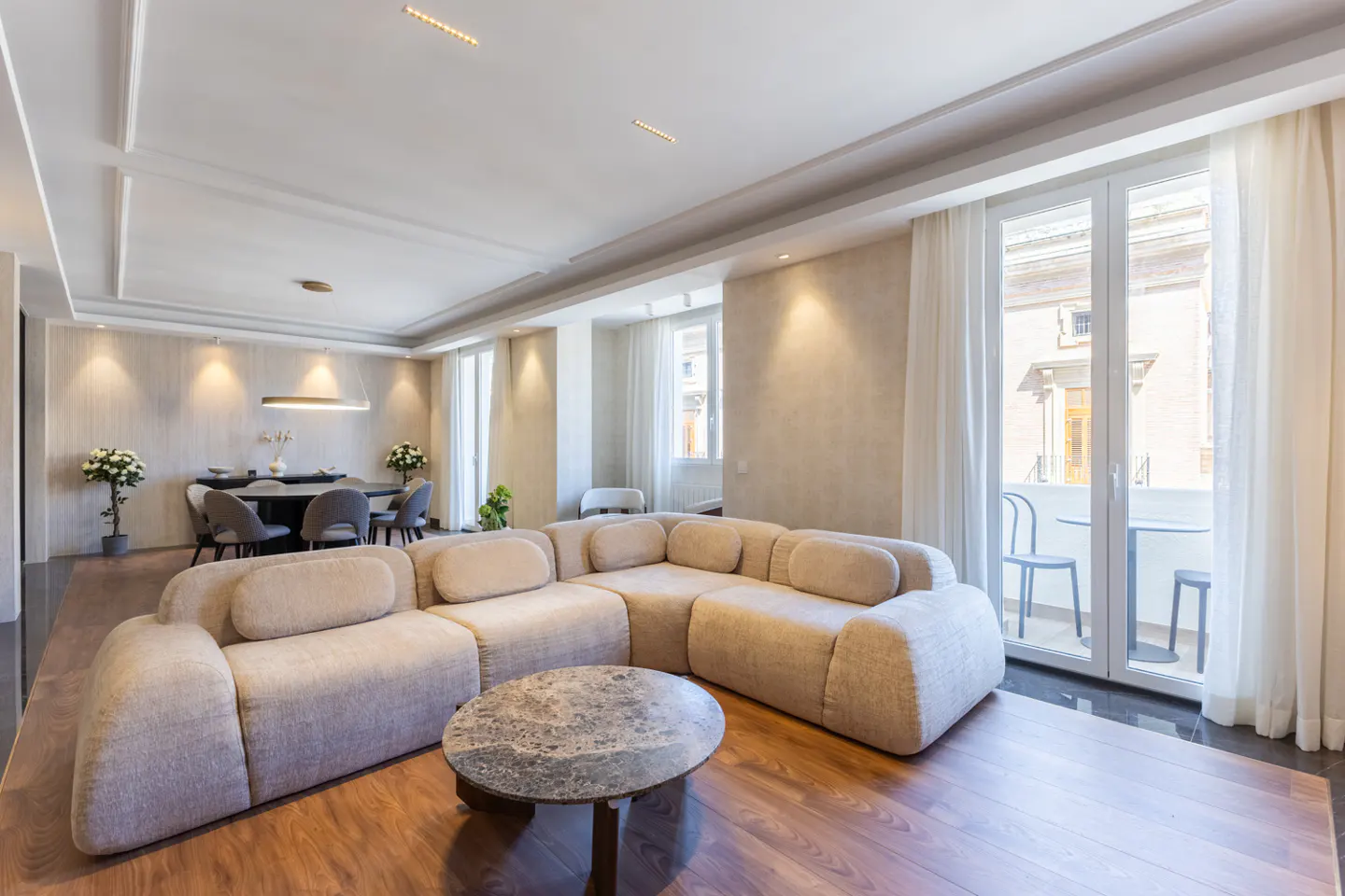 Modern living room with a beige sectional sofa, round marble coffee table, and a dining area with a balcony view.