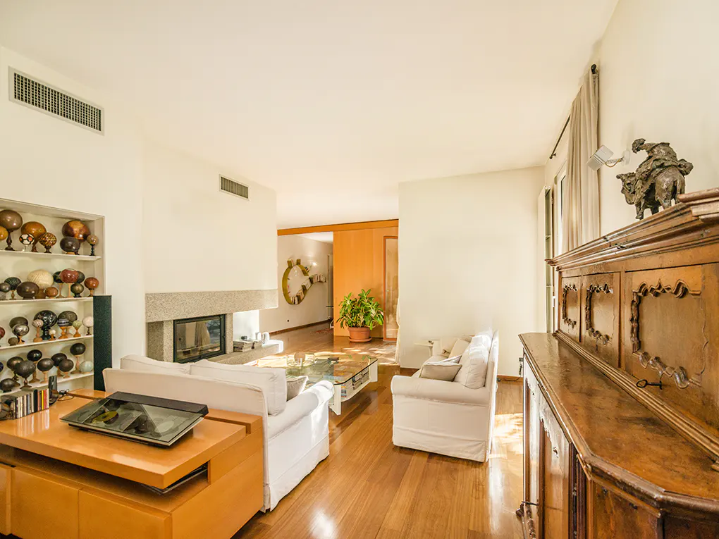 Bright living room with white sofas, a stone fireplace, and a wooden cabinet topped with a sculpture. Hardwood floors and natural light.