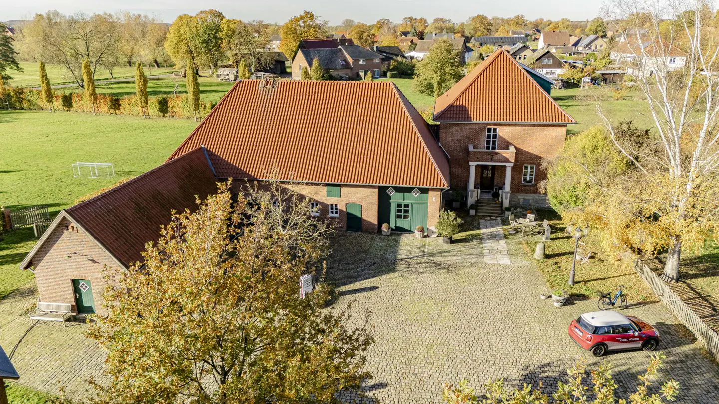 Aerial view of a red brick house with a red tile roof, green doors, and a cobblestone driveway with a red car.