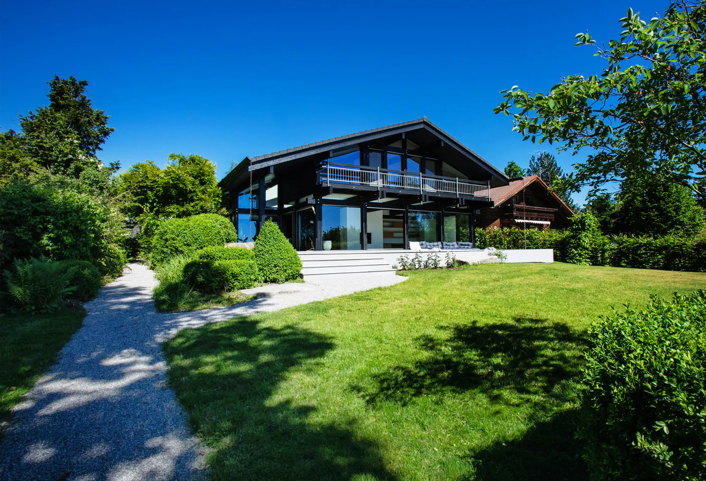 Two-story black house with large windows, a balcony, and a white patio, surrounded by green grass and trees under a blue sky.