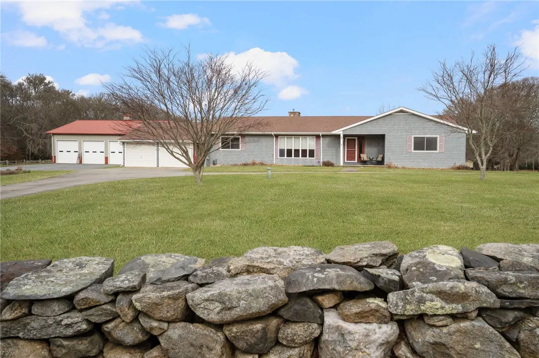 Gray house with red trim and a red-roofed garage, viewed over a stone wall on a green lawn.
