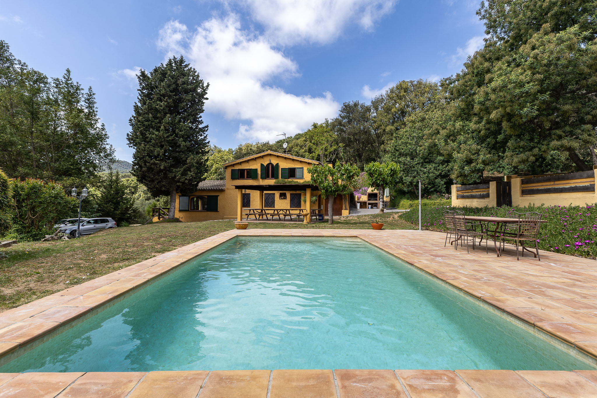 A rectangular pool with turquoise water in front of a yellow house with green shutters. Trees surround the property.