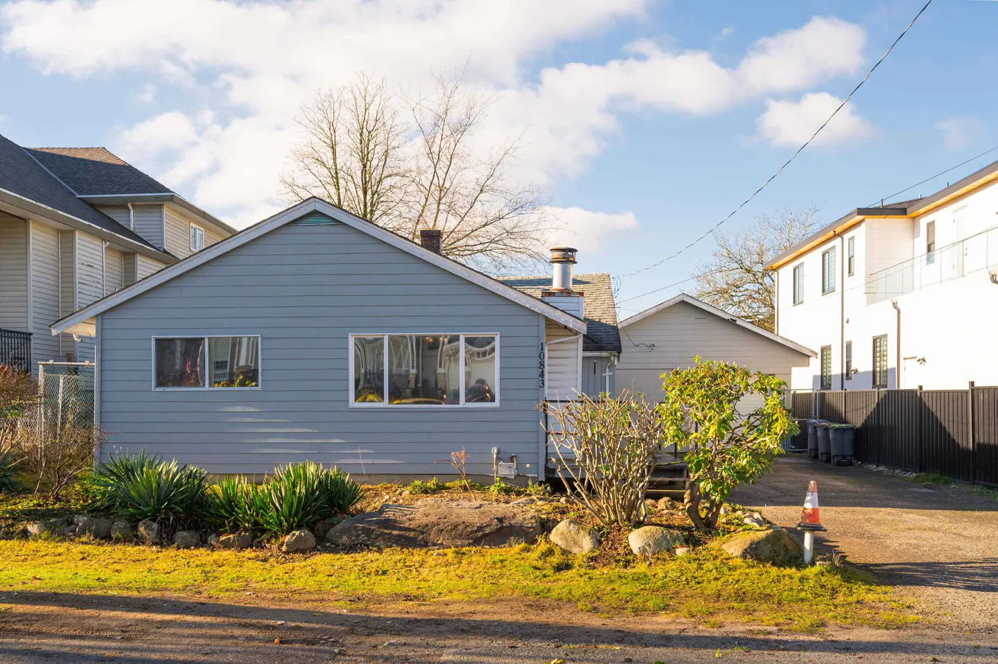 Single-story light blue house with a chimney, front yard landscaping, and a driveway leading to a detached garage.
