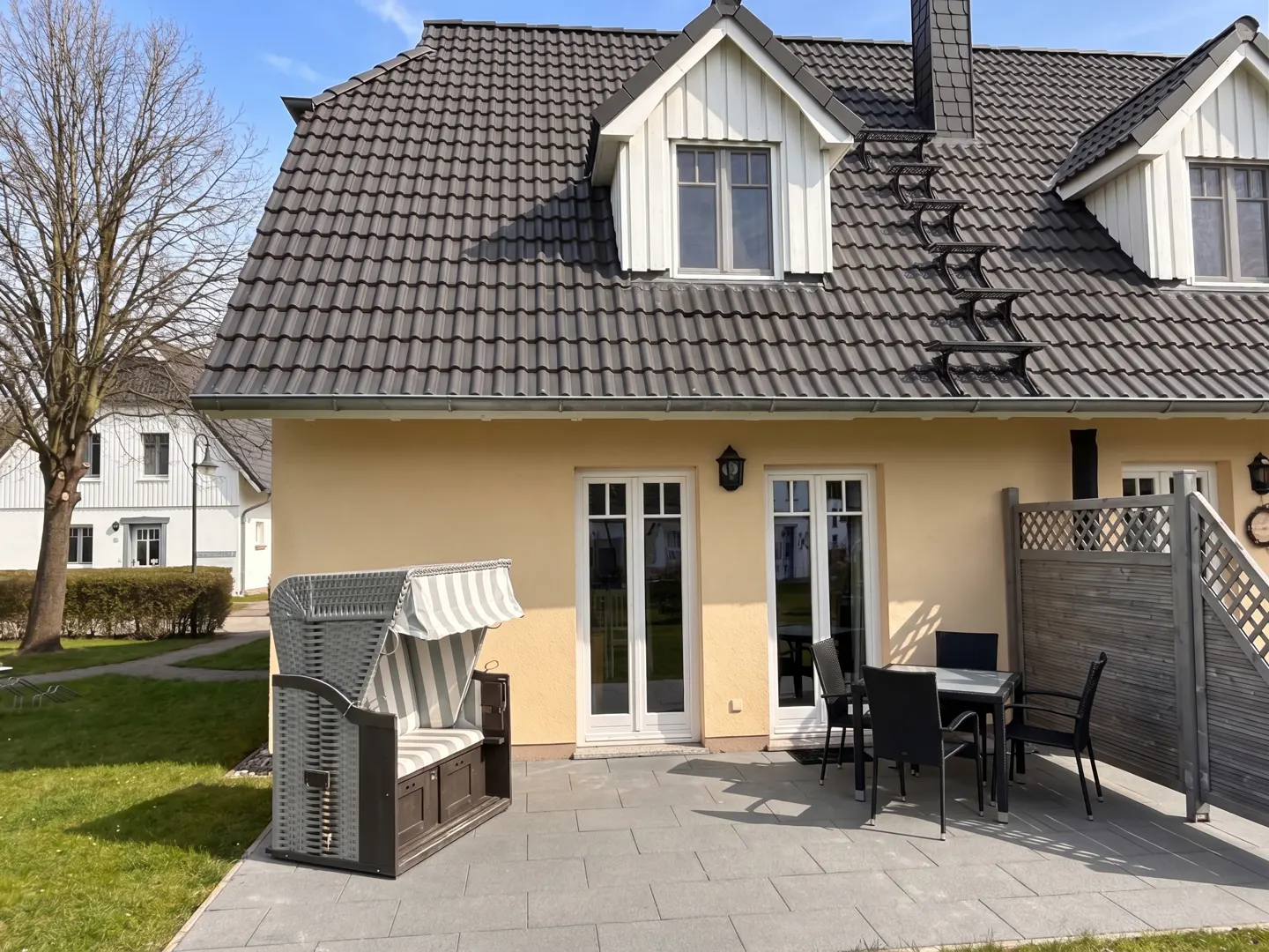 Exterior view of a yellow house with a gray tiled roof, dormer windows, and a roof ladder. A beach chair and patio furniture sit on a gray stone patio.