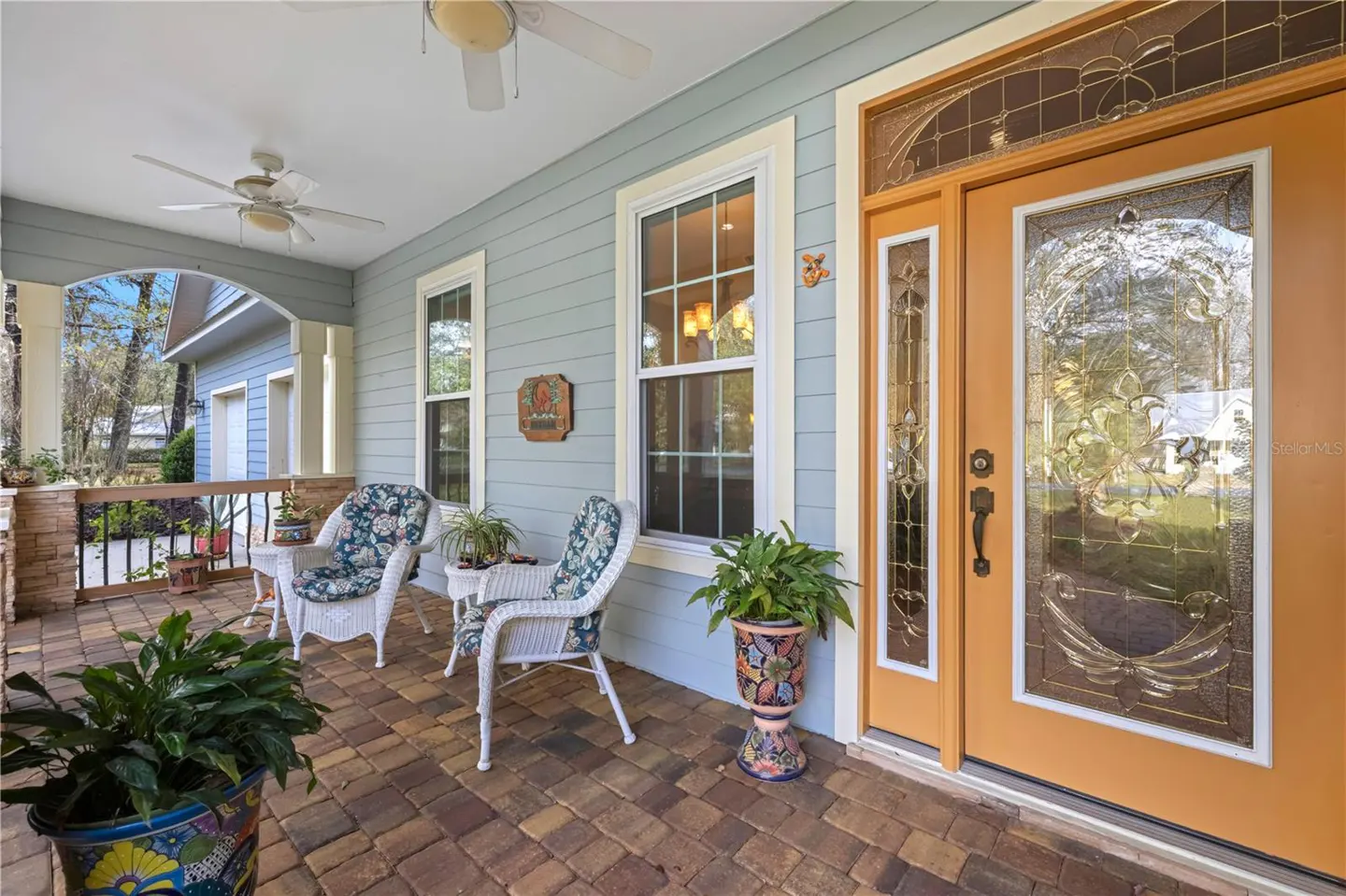 Covered porch with brick floor, wicker chairs with floral cushions, and a yellow front door with decorative glass.