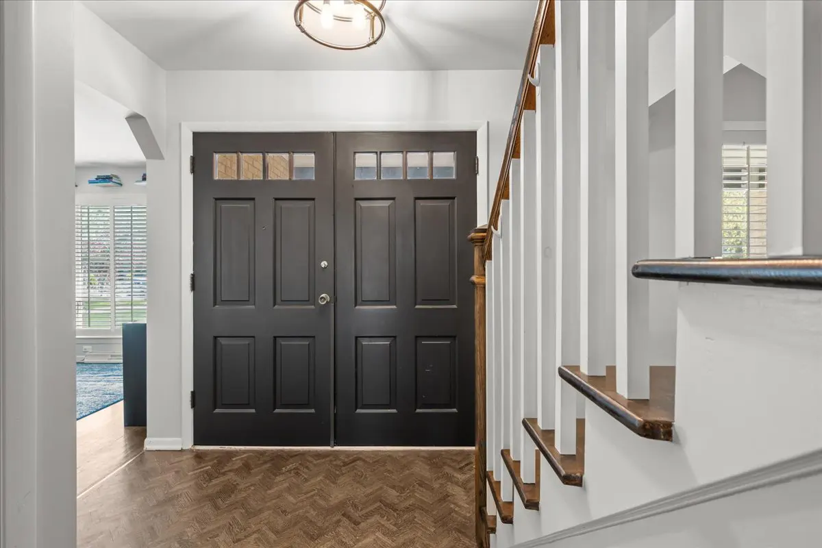 Entryway with dark double doors, herringbone wood floor, and white staircase with wood railing. A glimpse of a bright room is visible.
