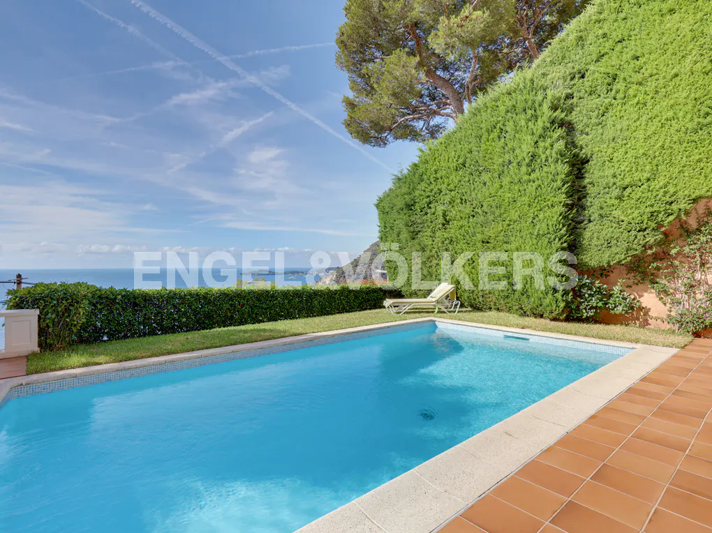 A rectangular swimming pool with blue water sits on a tiled patio next to a manicured hedge and a lounge chair by the sea.
