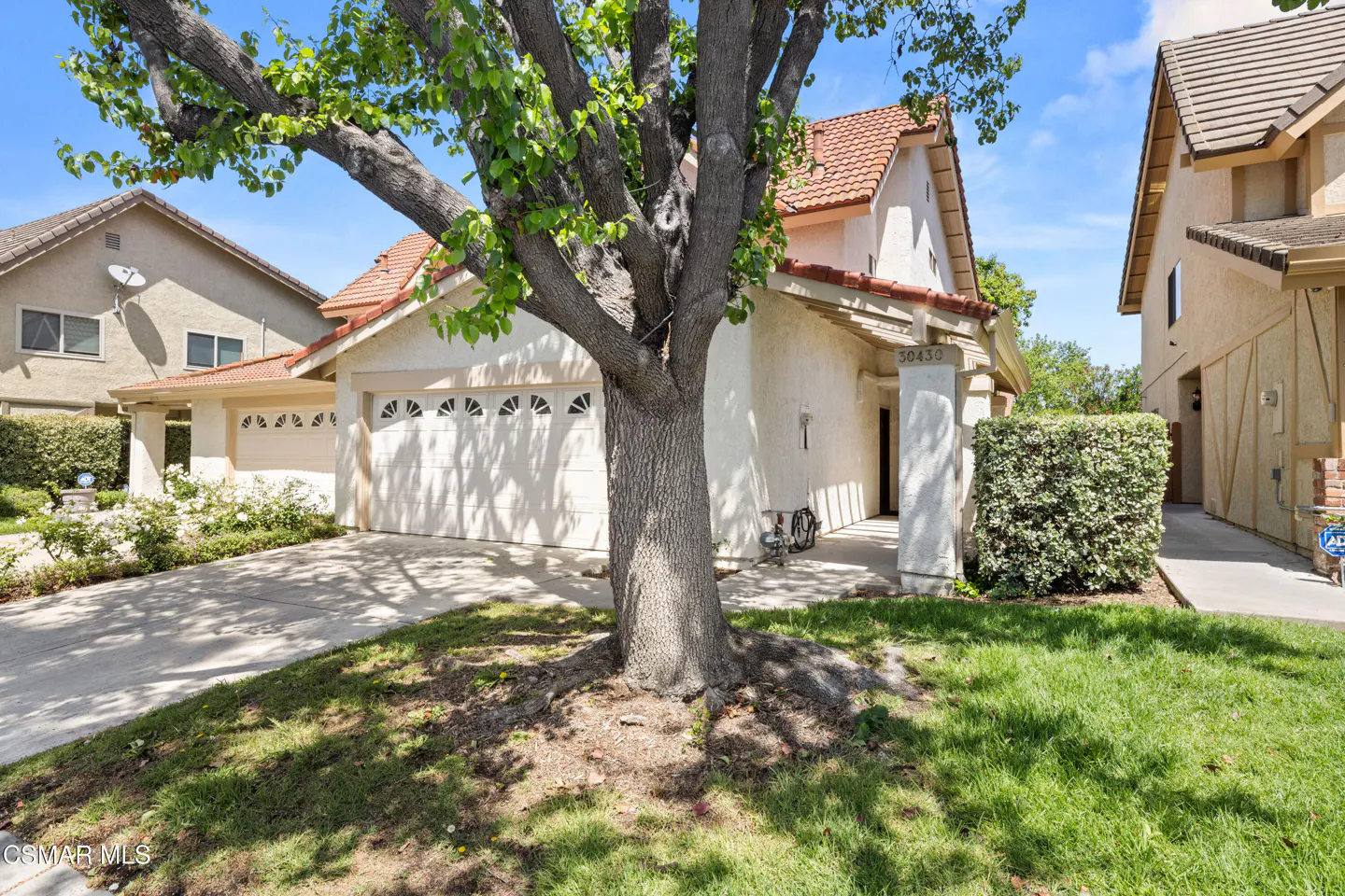 A beige single-story house with a red tile roof and a white garage door is partially obscured by a large tree.