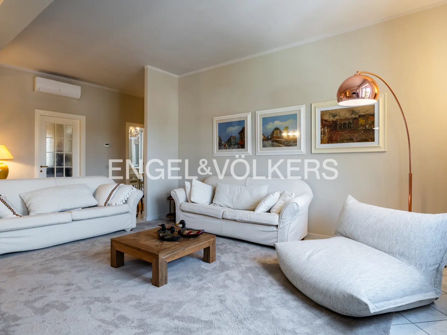 Living room with white sofas, a wooden coffee table with duck figurines, and a copper floor lamp. Three framed paintings hang on the wall.