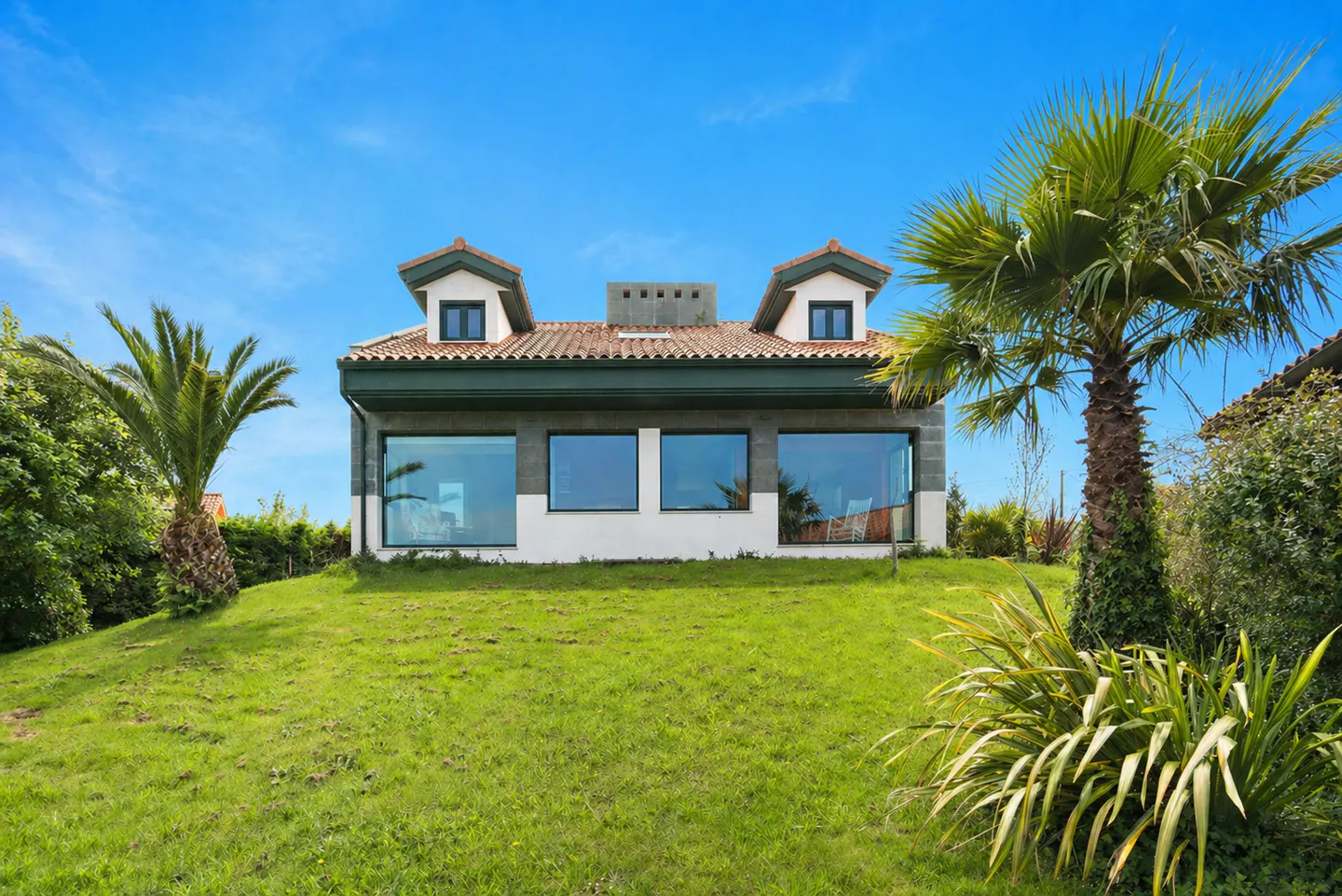 A two-story house with a green lawn, palm trees, and a blue sky. The house has a red tile roof and large windows.