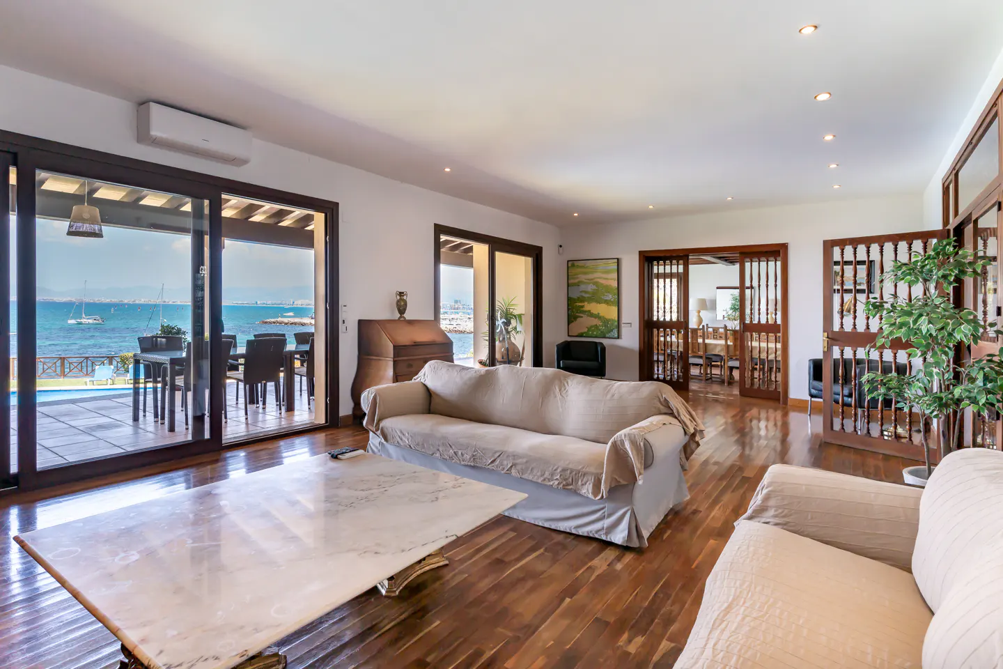 Bright living room with wood floors, beige sofas, and a marble coffee table. Sliding glass doors open to a patio with ocean views.