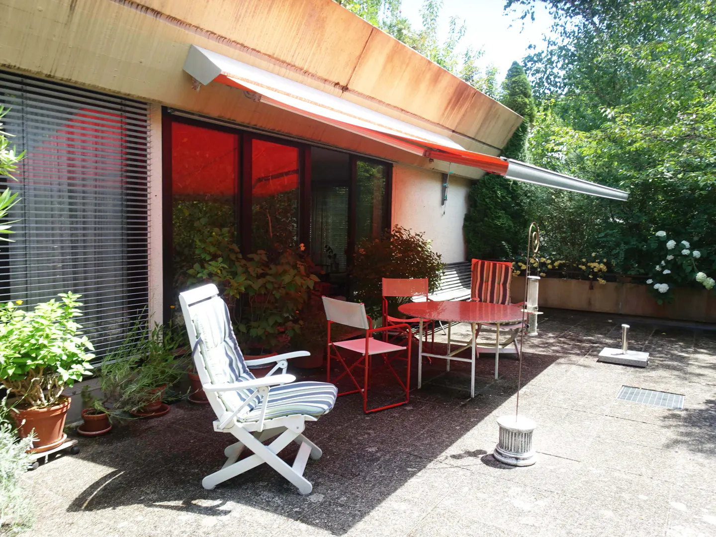 Outdoor patio with red table and chairs, white folding chair, and awning. Lush greenery in the background.
