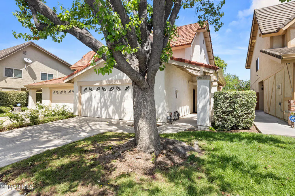 Beige single-story house with a red tile roof and a two-car garage, framed by a large tree and green lawn.