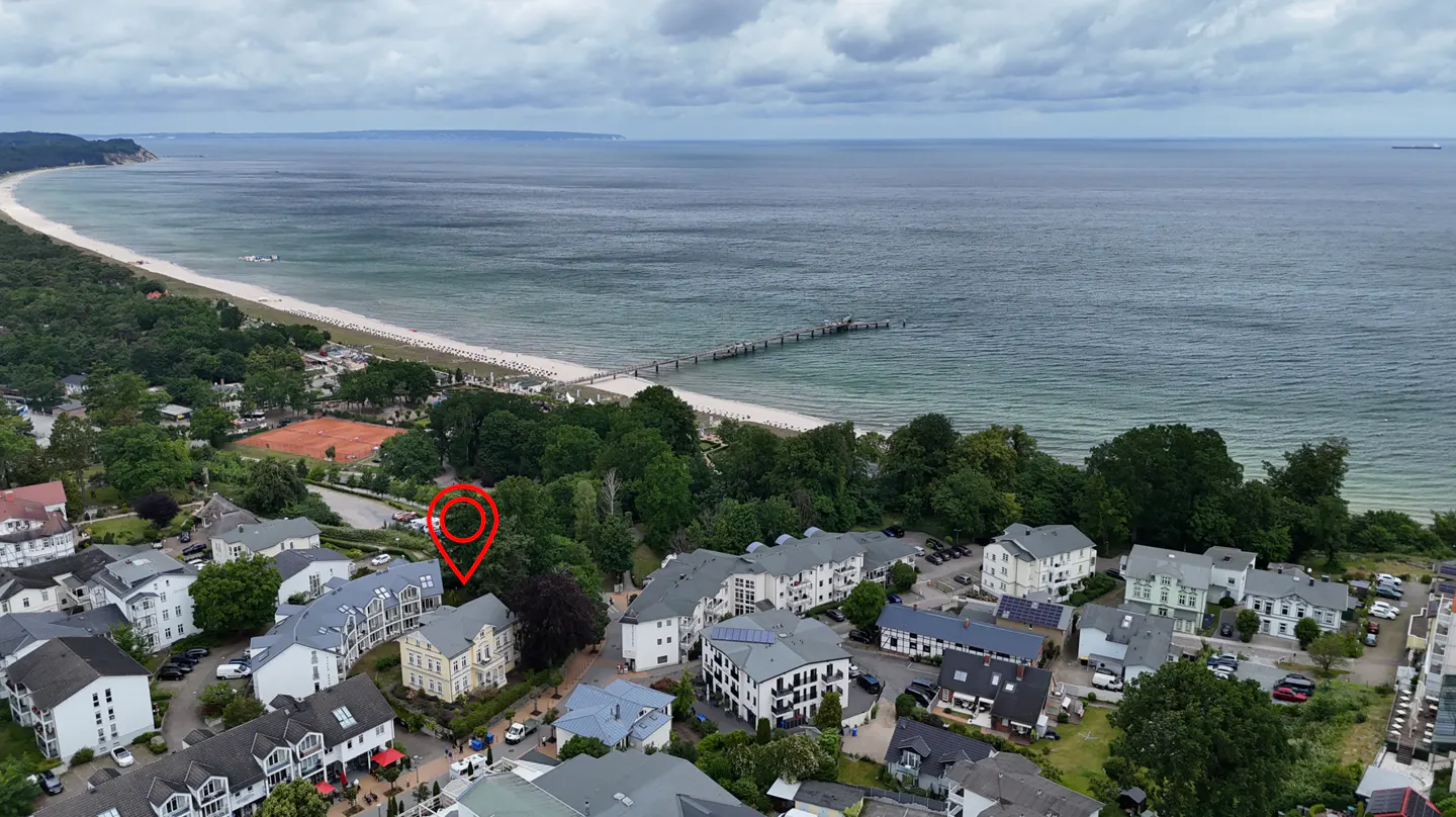 Aerial view of a coastal town with a red location pin marking a yellow building near a sandy beach and a pier.