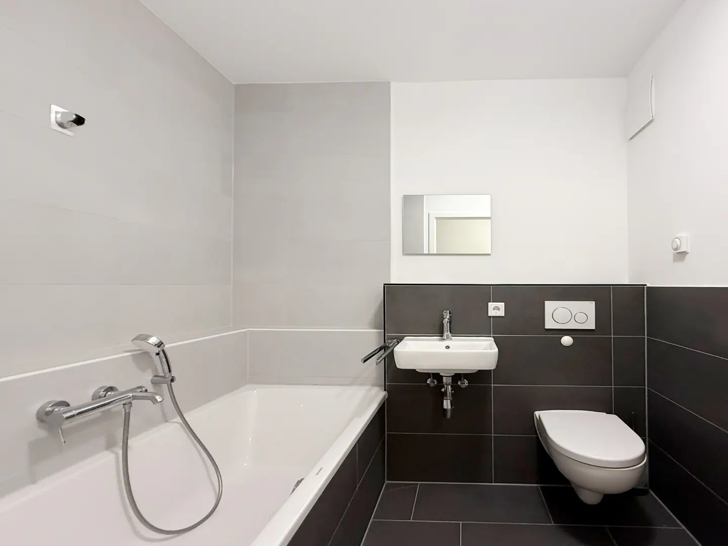 Modern bathroom with white tub, sink, and toilet against gray and dark tile walls. A mirror hangs above the sink.