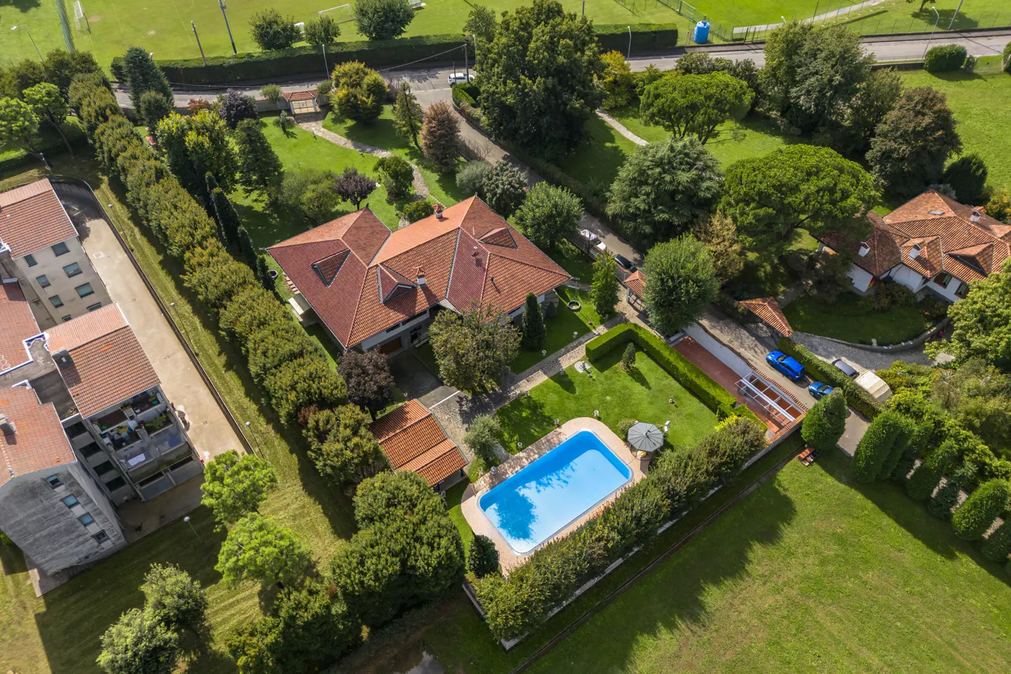 Aerial view of a large house with a red tile roof, a blue swimming pool, and green lawns surrounded by trees and hedges.