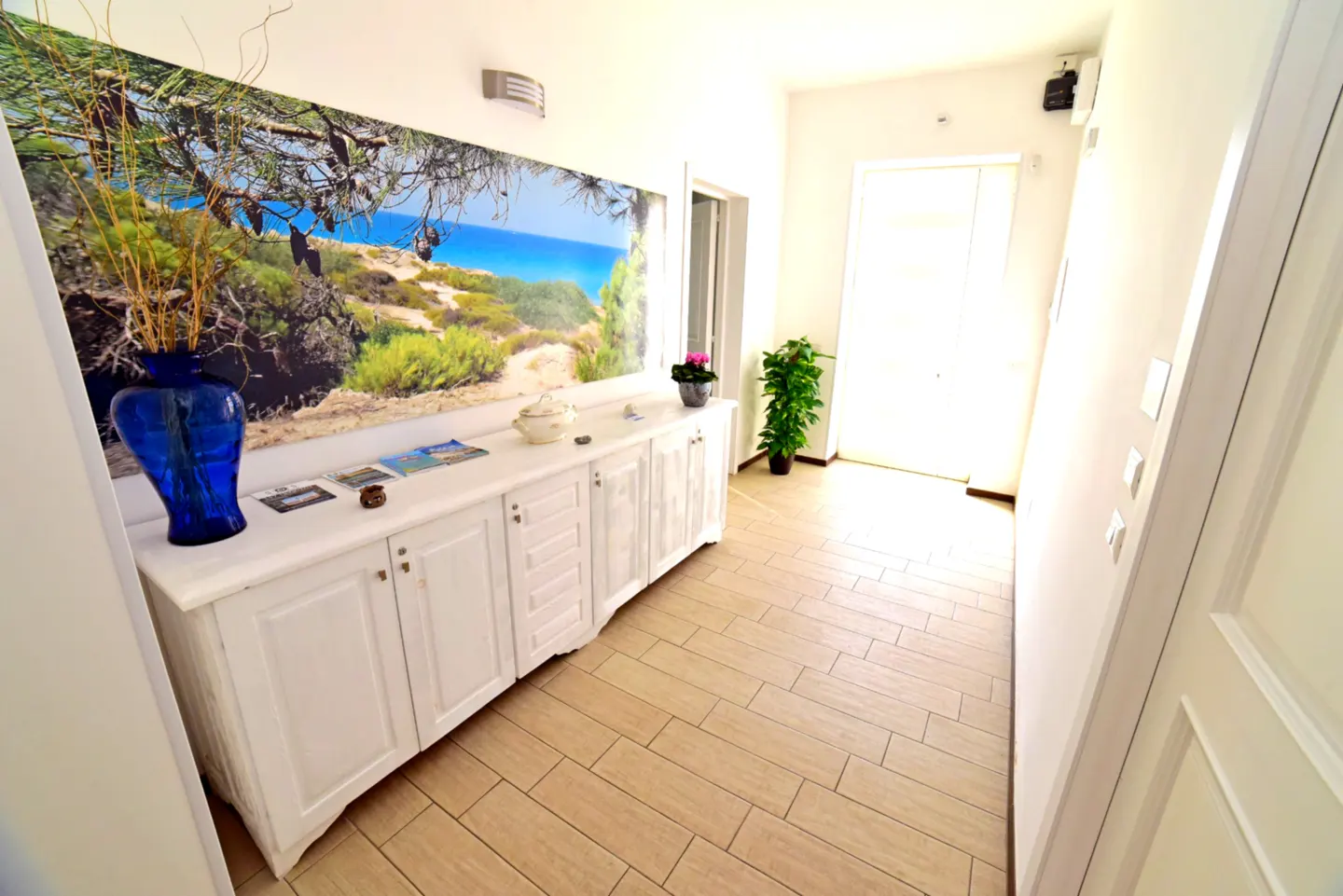 Bright hallway with beige tile floor, white walls, and a white cabinet. A blue vase sits on the cabinet under a large beach scene print. A door is visible at the end of the hall.