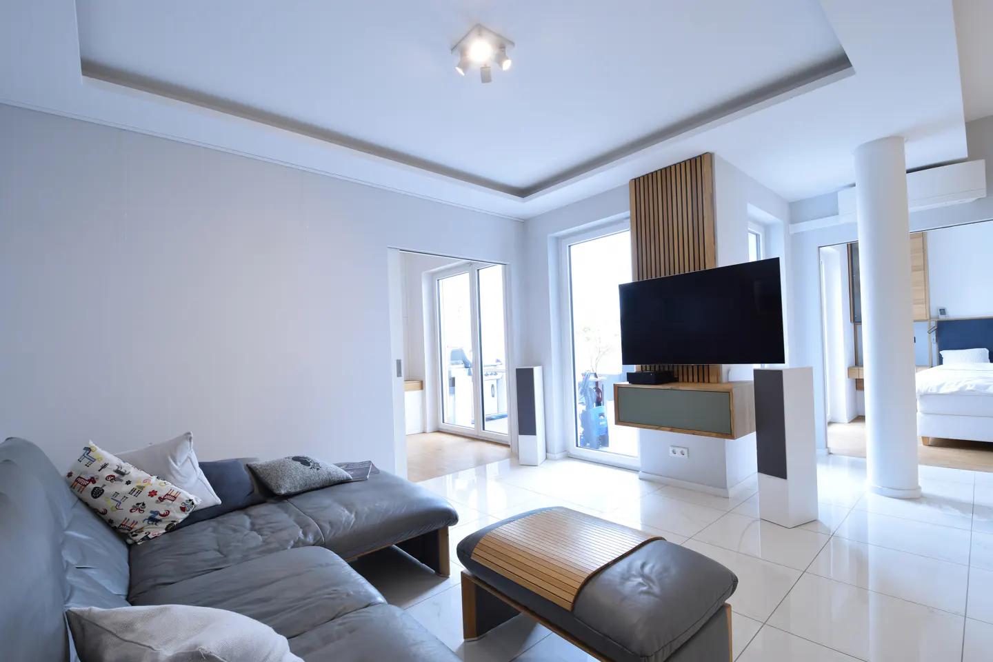 Modern living room with gray sectional sofa, ottoman, TV, and speakers. White walls and tile floors create a bright, airy space.