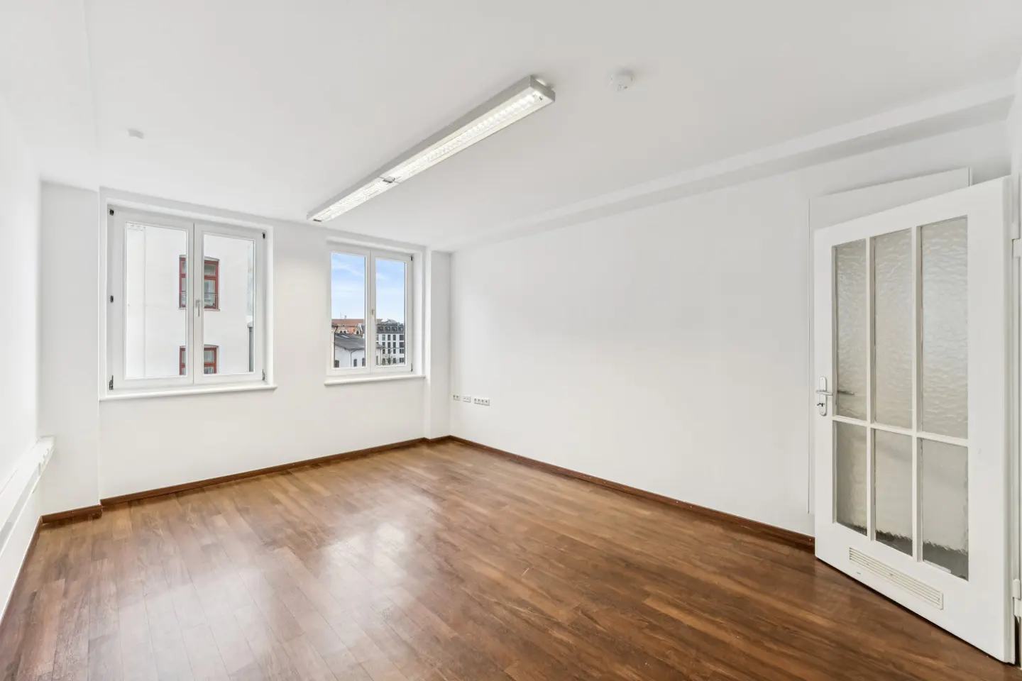 Bright, empty room with hardwood floors, white walls, and two windows. A white door with glass panes is partially open.