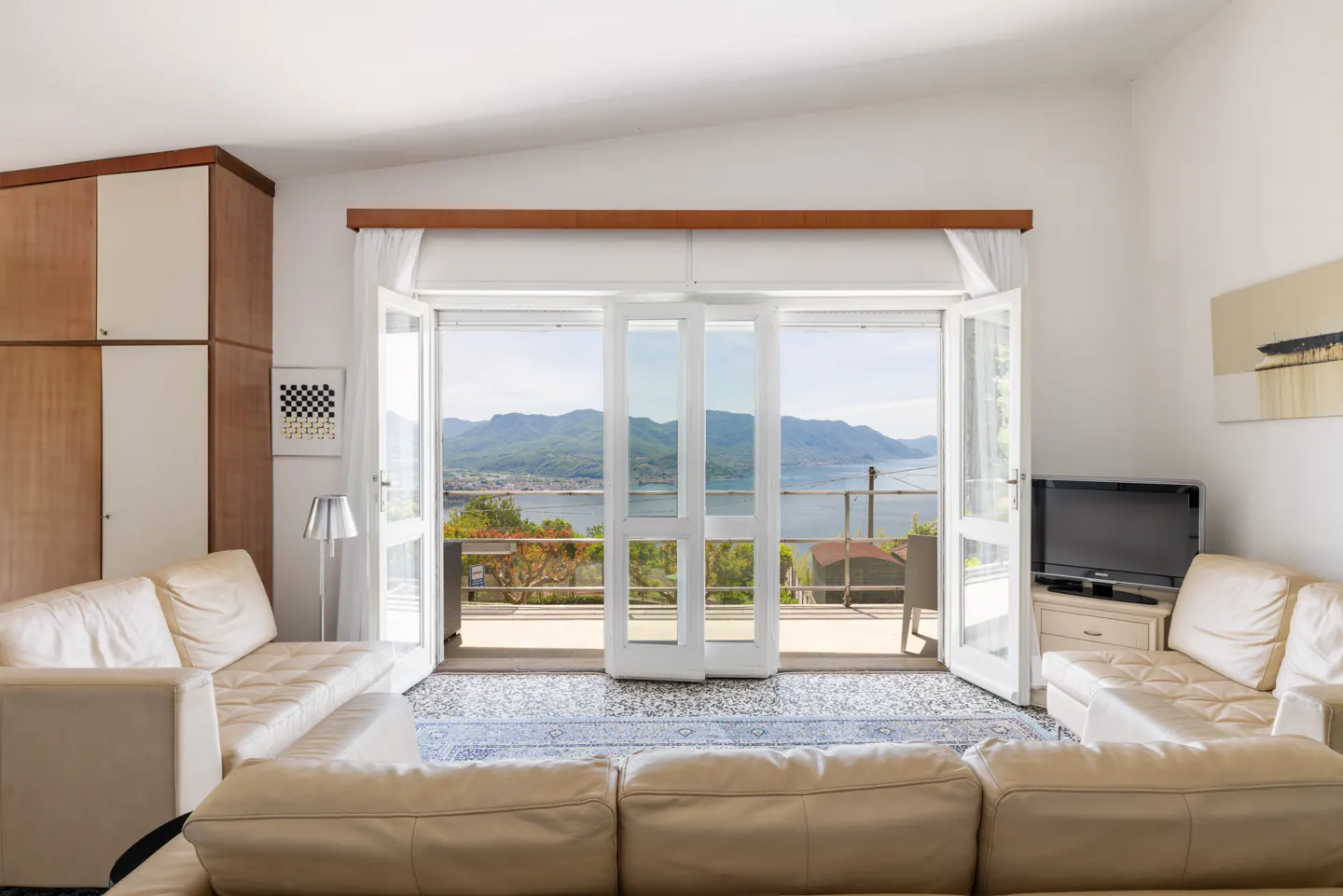 Living room with white sofas, a TV, and a balcony with a view of mountains and water.