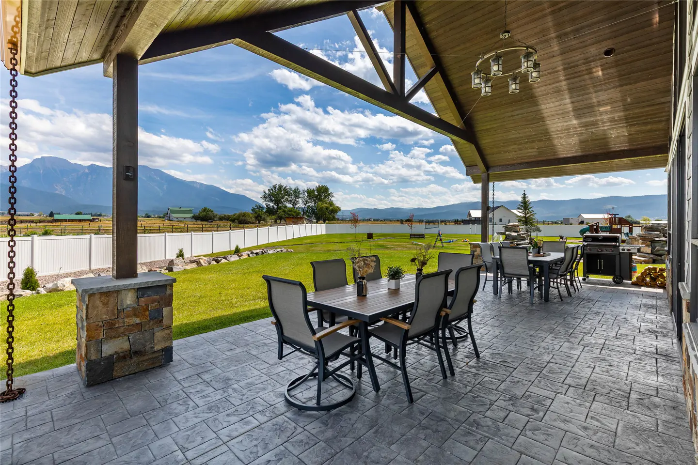 Covered stone patio with dining set, overlooking a grassy backyard, white fence, and distant mountain range under blue sky.