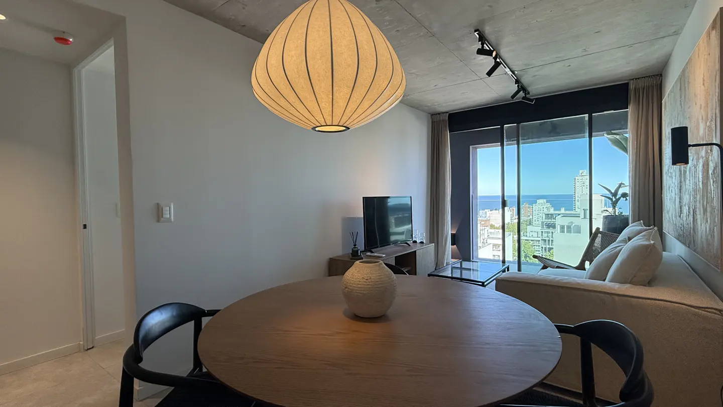 A modern apartment interior with a round wooden table, black chairs, and a large window overlooking the ocean. A beige sofa and TV are visible in the background.