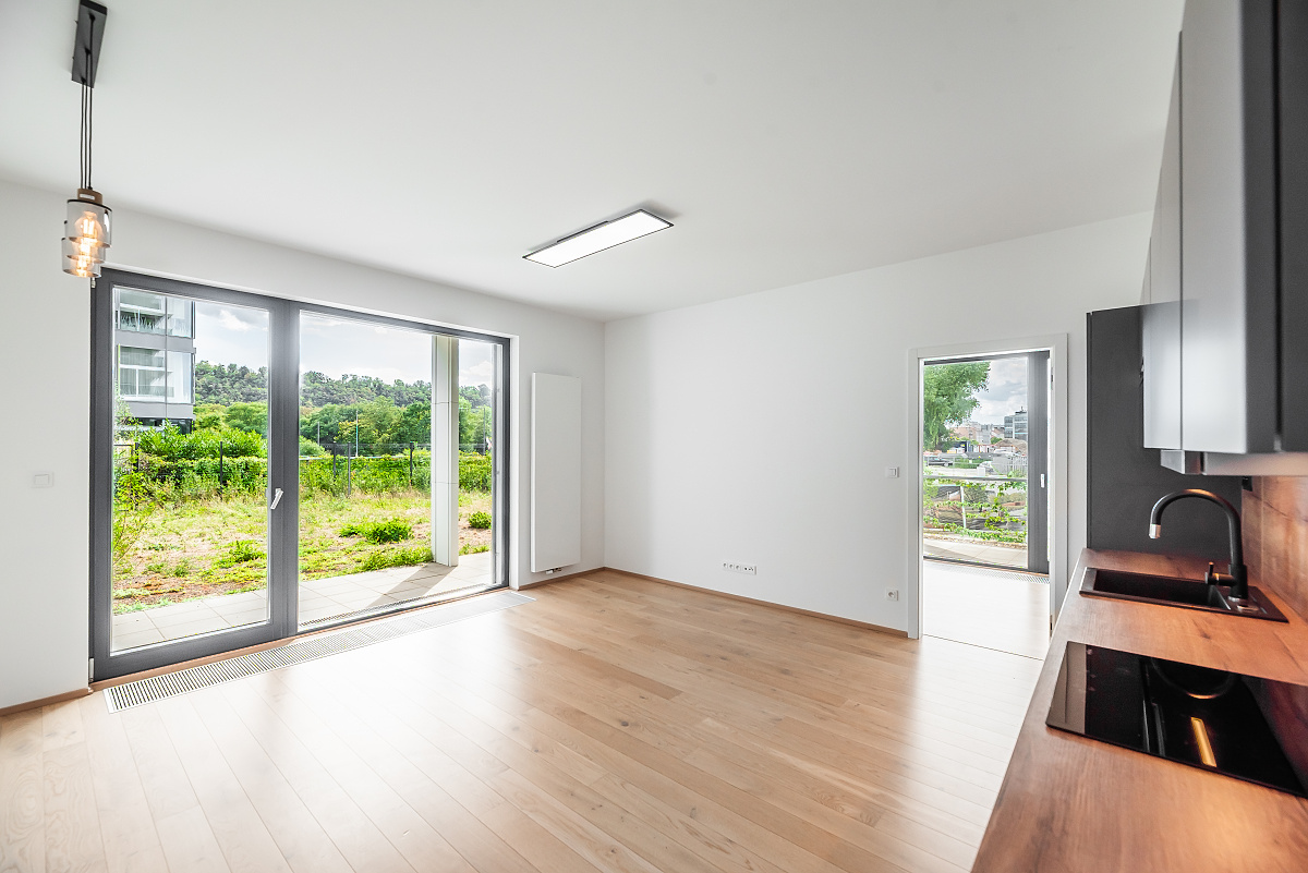 Modern open-plan room with light wood floors, white walls, a kitchen area, and large glass doors opening to a green yard. [AI-generated text]