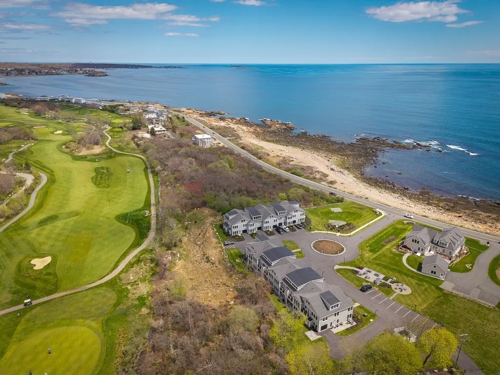 Aerial view of gray townhouses near a green golf course and blue ocean under a partly cloudy sky.