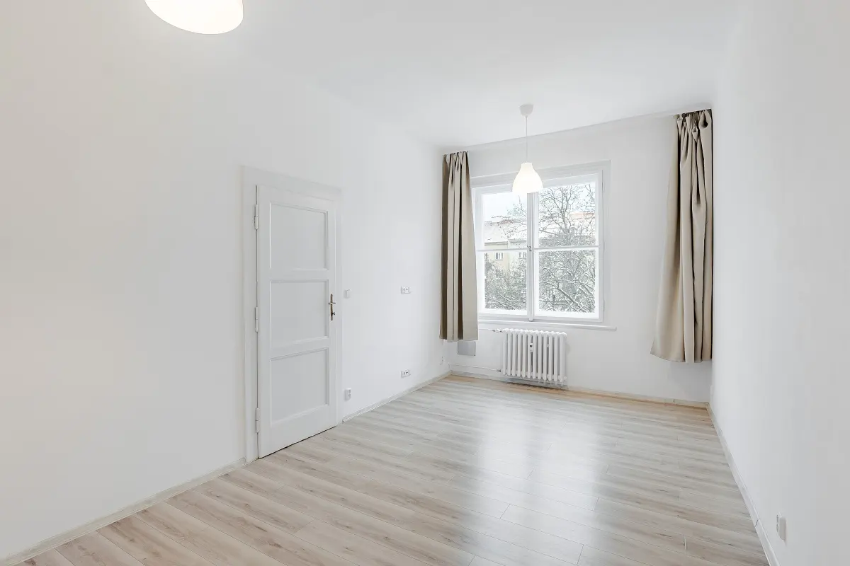 Bright, empty room with white walls, light wood floors, and a window with beige curtains. A white door is on the left.