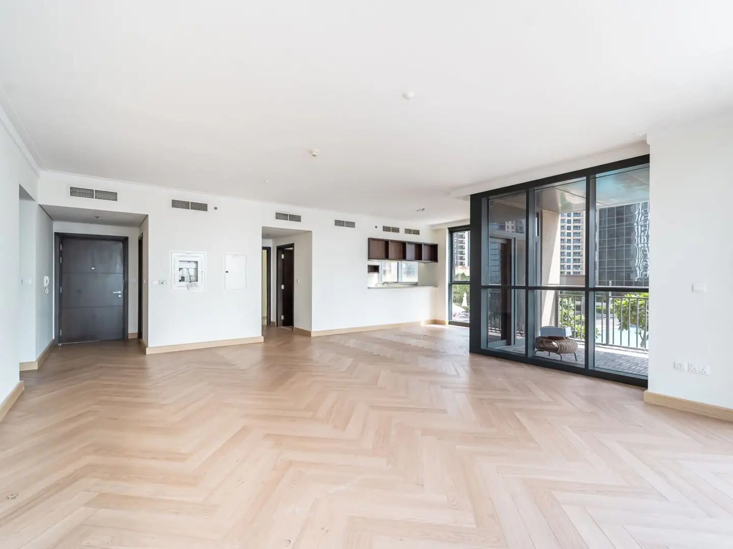 Bright, empty apartment with herringbone wood floors, white walls, and a large window to a balcony with city views.