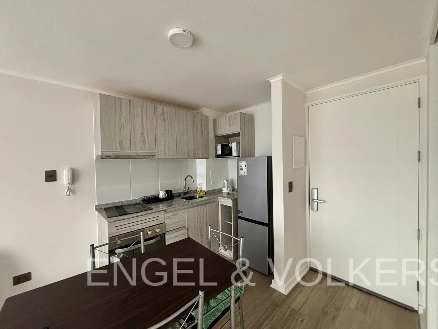A bright kitchen with wood cabinets, stainless steel appliances, and a dark wood table with green chairs. A white door is on the right.