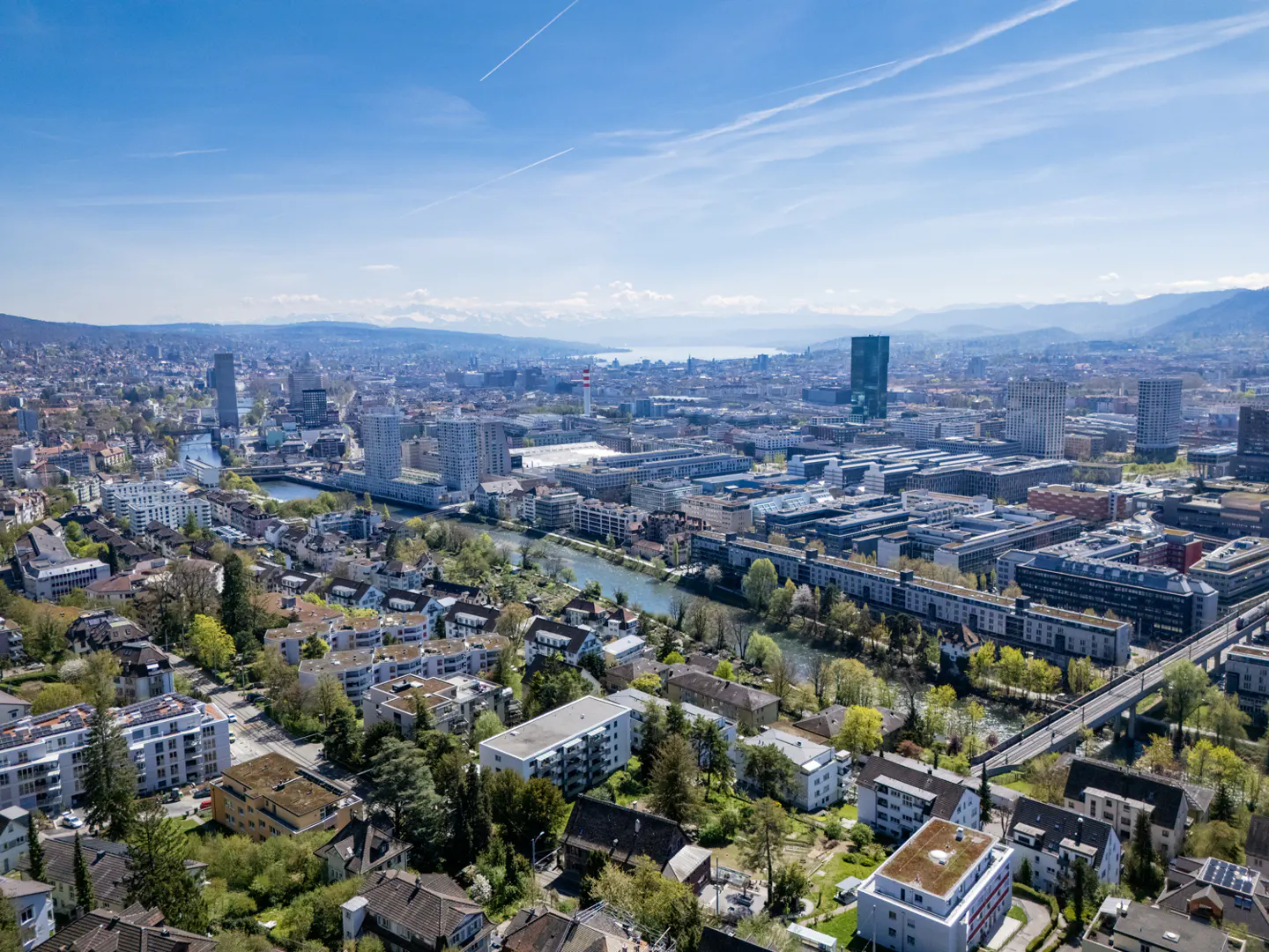 Aerial view of Zurich, Switzerland, showing a river winding through the city, with buildings, trees, and mountains in the background under a blue sky.