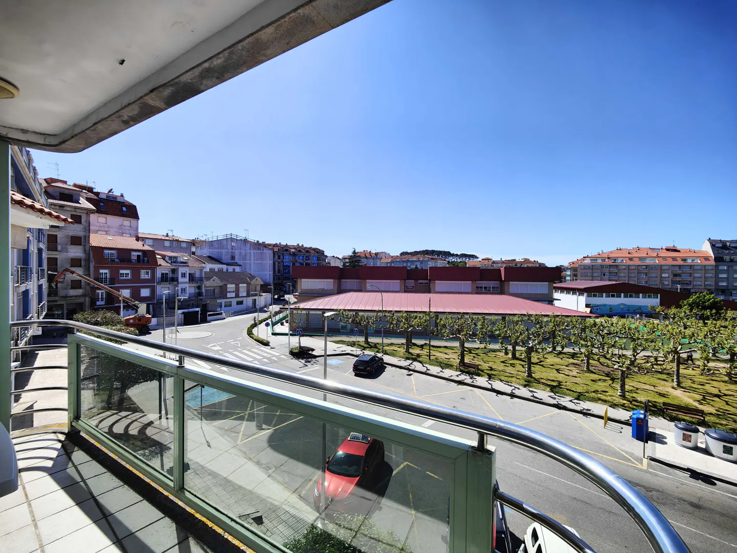 View from a balcony with glass railings overlooking a street with cars, buildings, and a park under a clear blue sky.