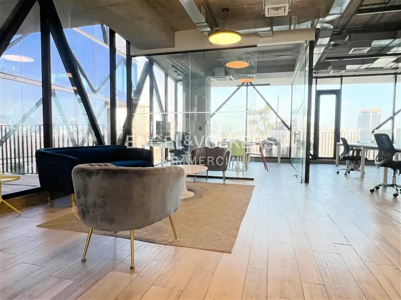 Open office space with blue sofa, gray chairs, and desks. Large windows offer a city view. Modern lighting fixtures hang from the ceiling.