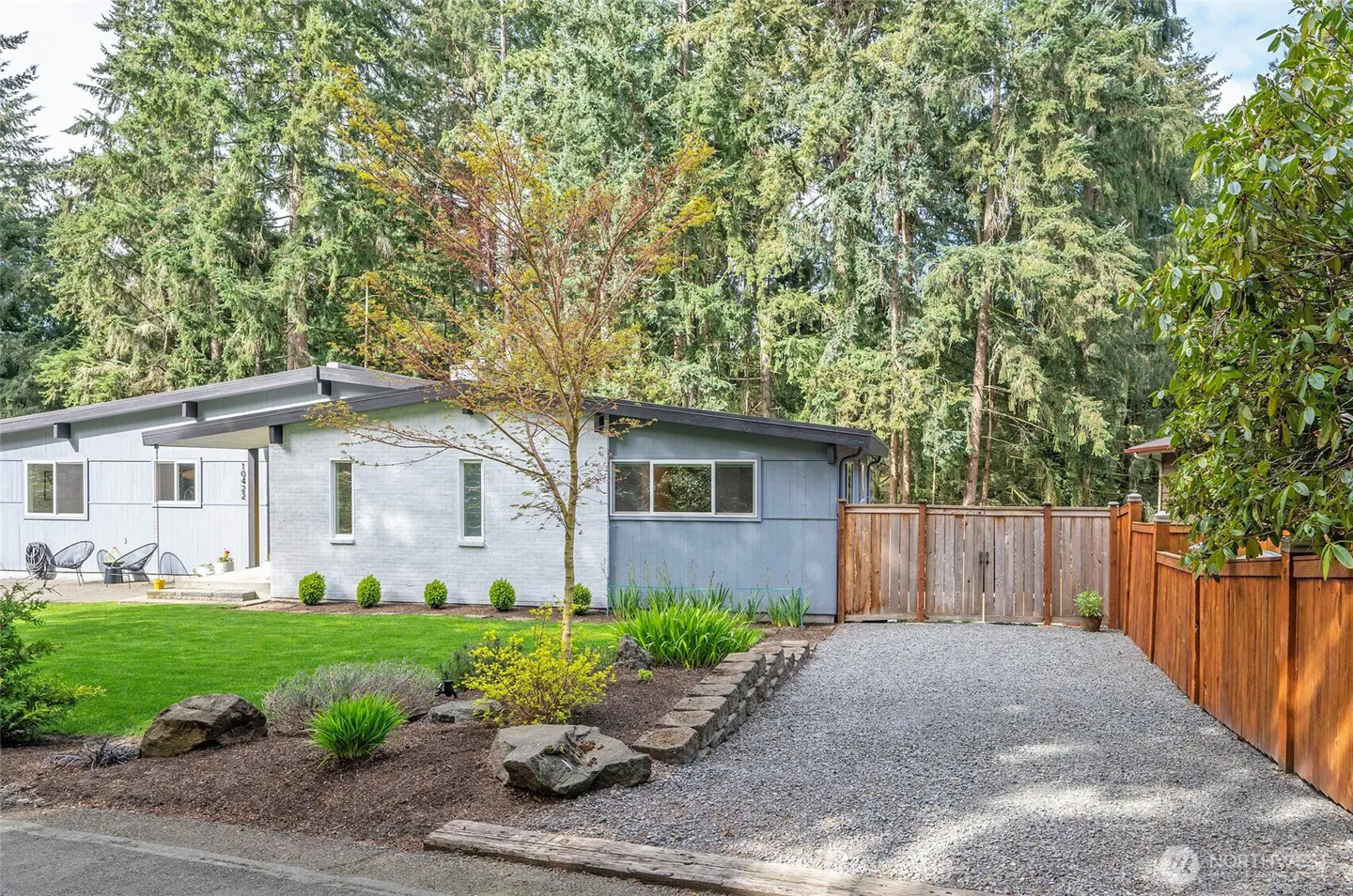 Exterior view of a light blue single-story house with a gray roof, green lawn, and gravel driveway. Tall trees are in the background.