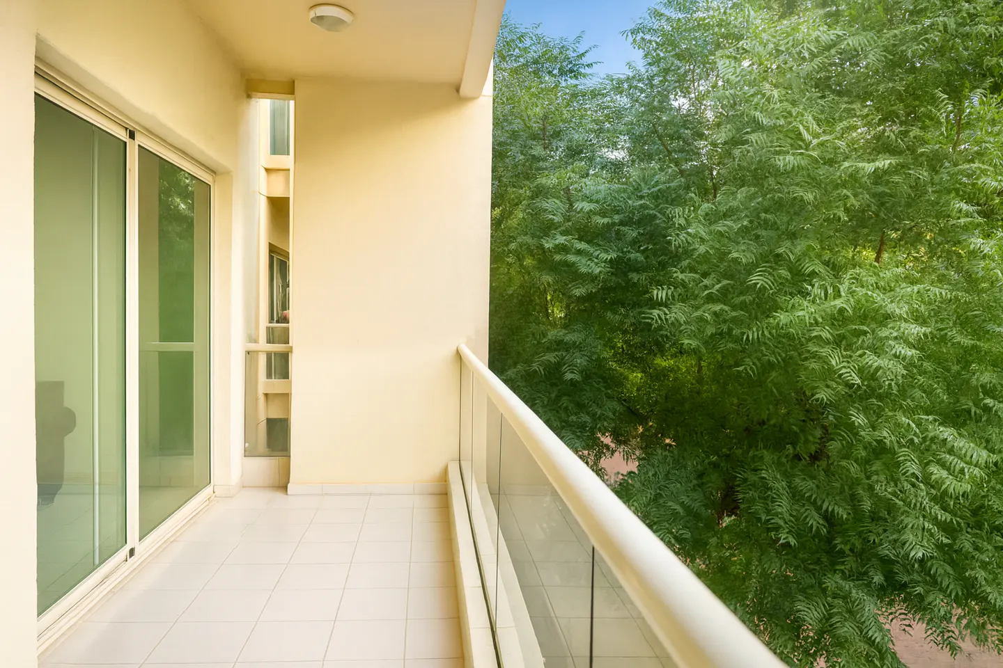 Balcony with white tile floor, glass railing, and sliding glass doors. Lush green trees are visible in the background.