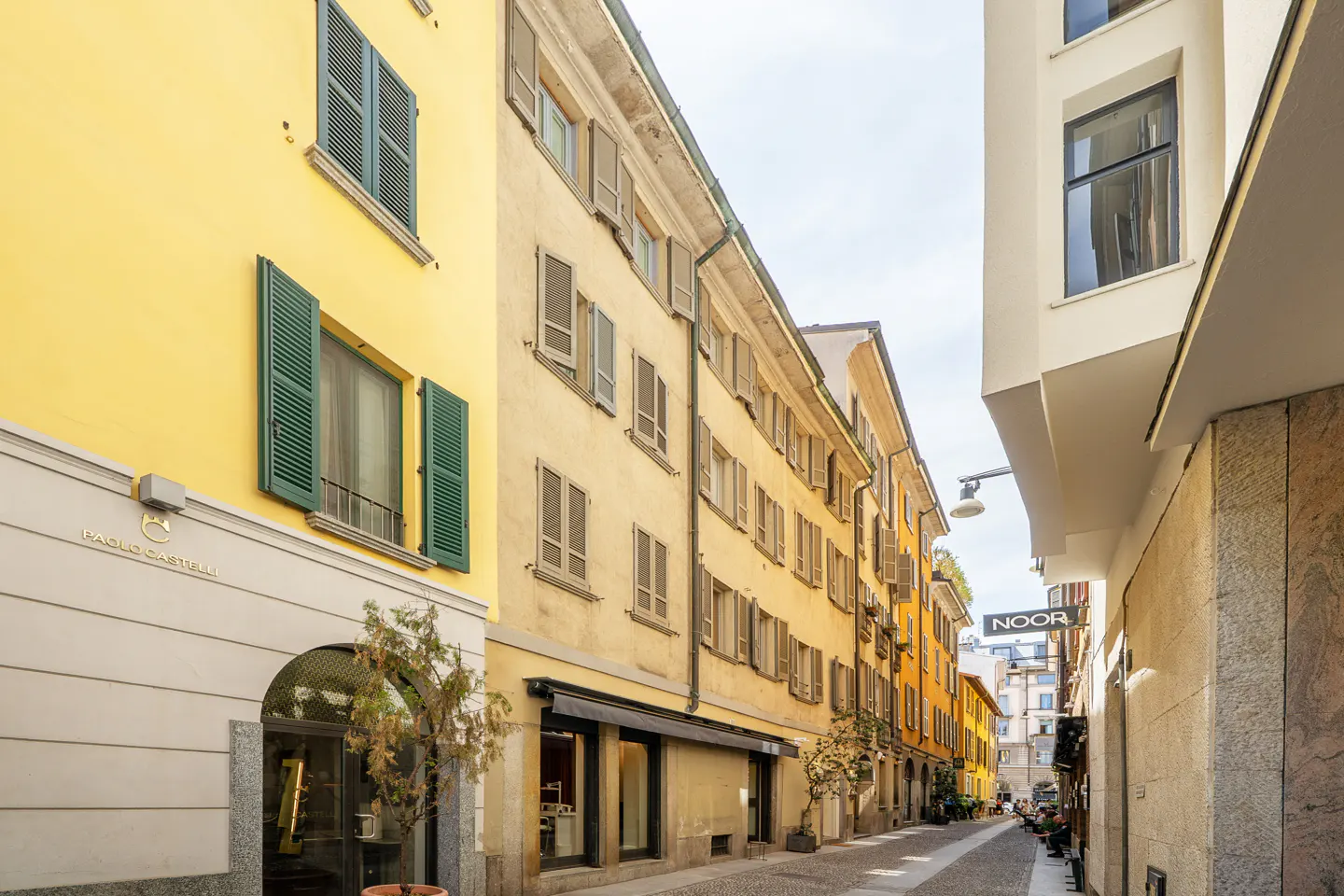 A narrow European street lined with tall, yellow and beige historic buildings featuring green shutters and storefronts.