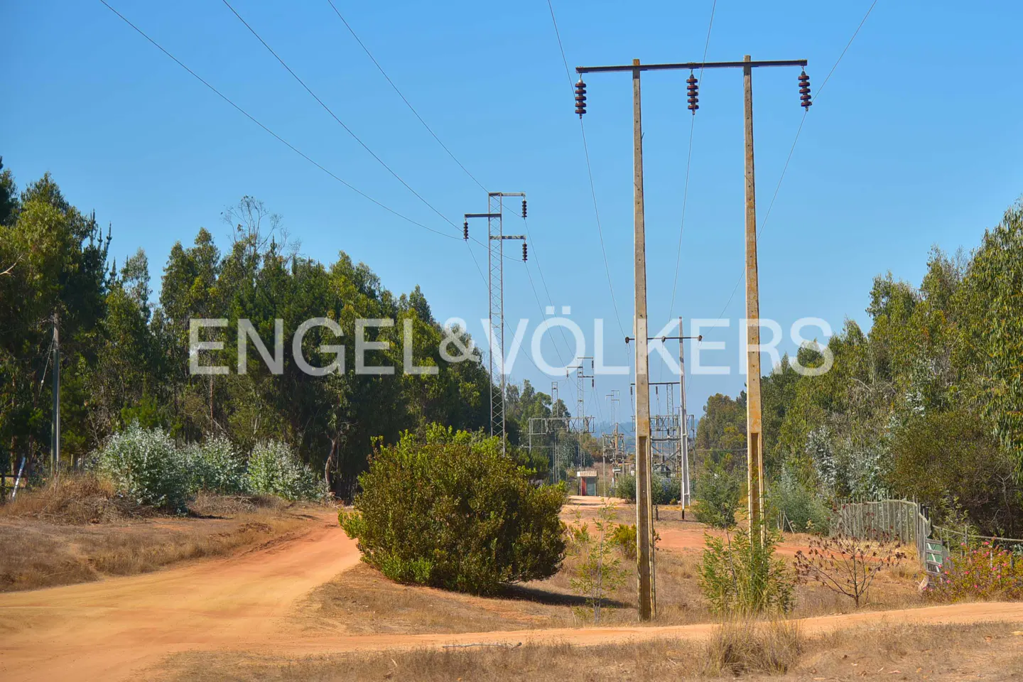 Dirt road with power lines and poles under a blue sky, surrounded by green trees and bushes. Engel & Völkers logo is visible.