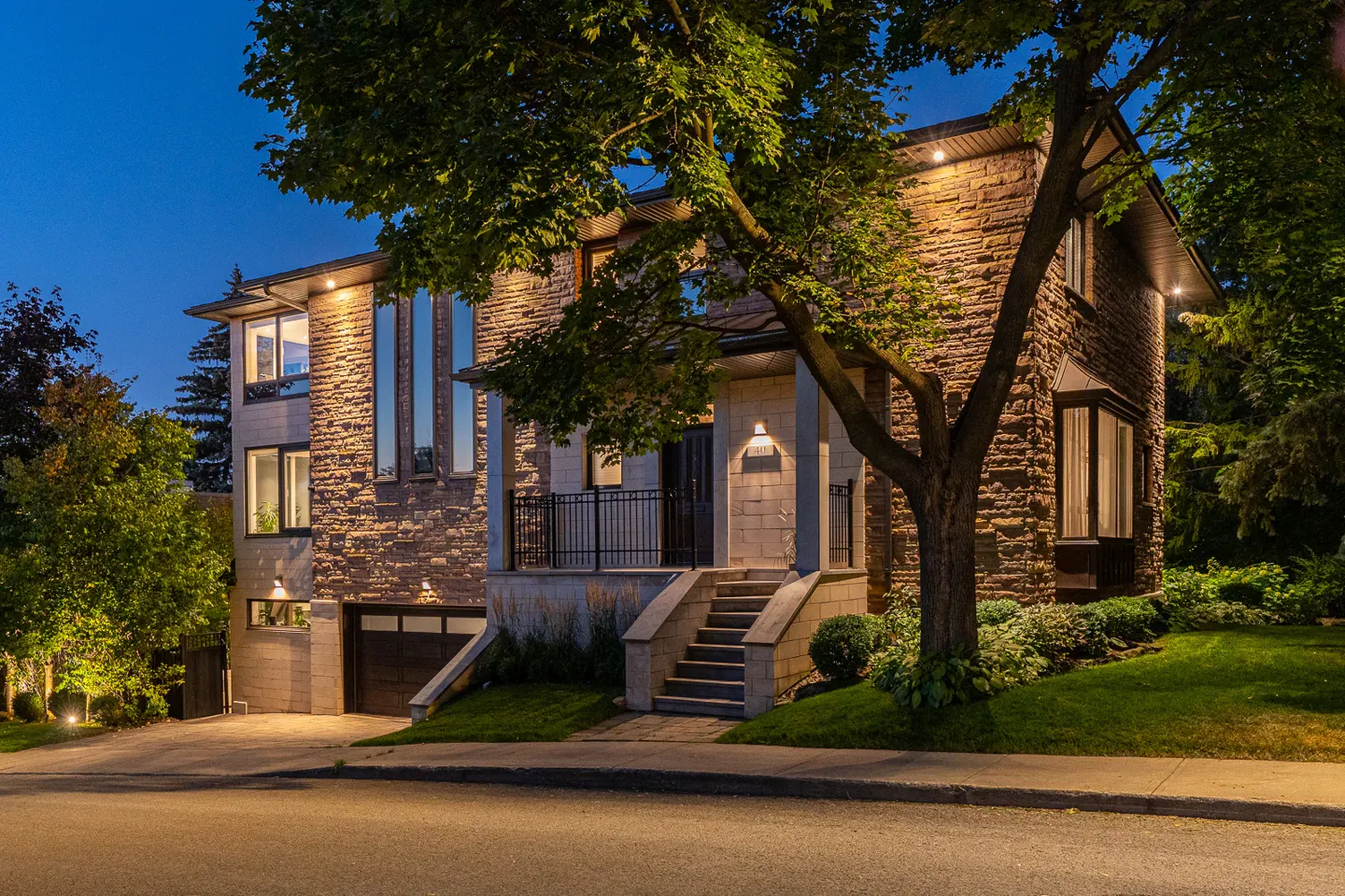 A two-story stone house at dusk with a tree in the foreground. The house has a garage and a porch with stairs.