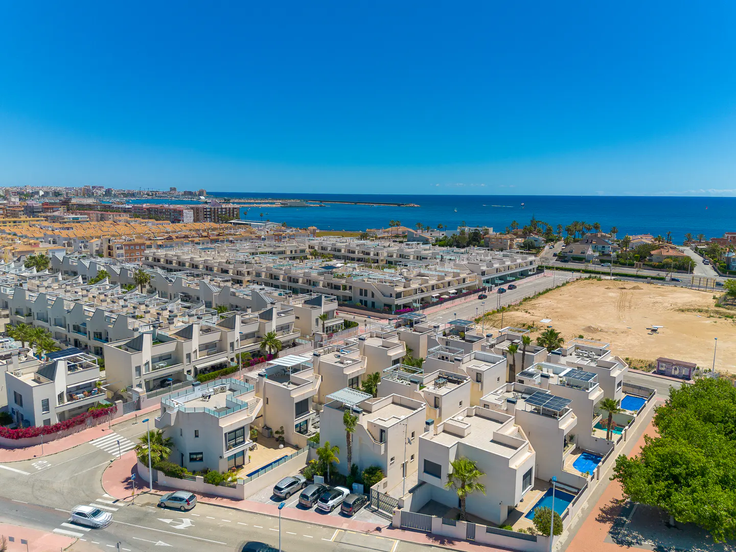 Aerial view of a residential area with white houses, blue pools, and the ocean in the background under a clear blue sky.
