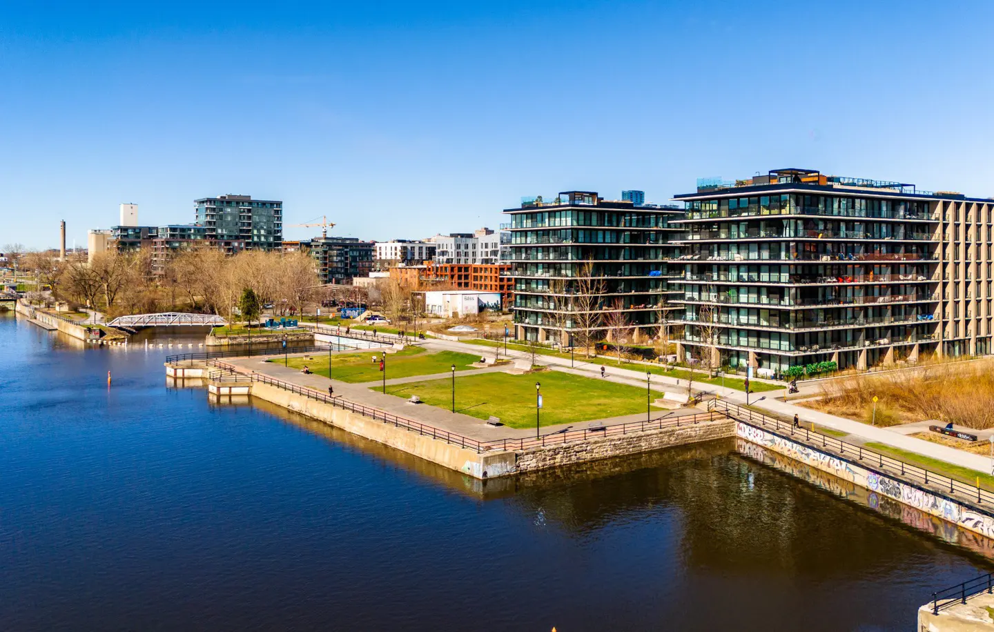 Waterfront condos with balconies overlook a green park and blue canal under a clear sky.