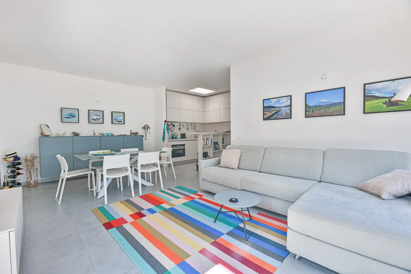 Bright living room with a gray sectional sofa, colorful striped rug, and white dining set. Kitchen is visible in the background.