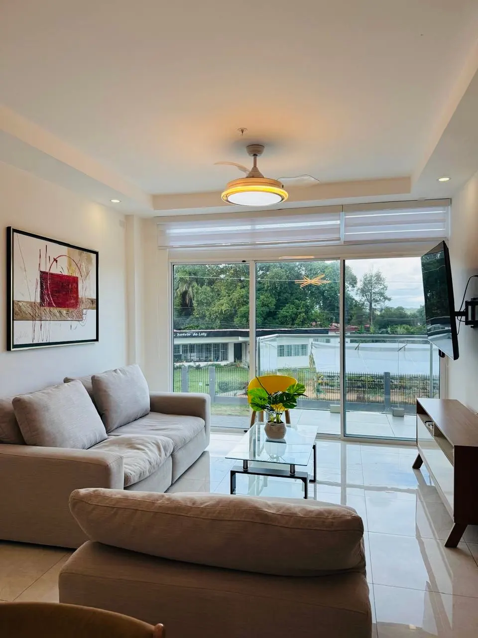 Bright living room with a beige sofa, glass table, and a yellow chair. Large windows offer a view of lush greenery. Modern art hangs on the wall.