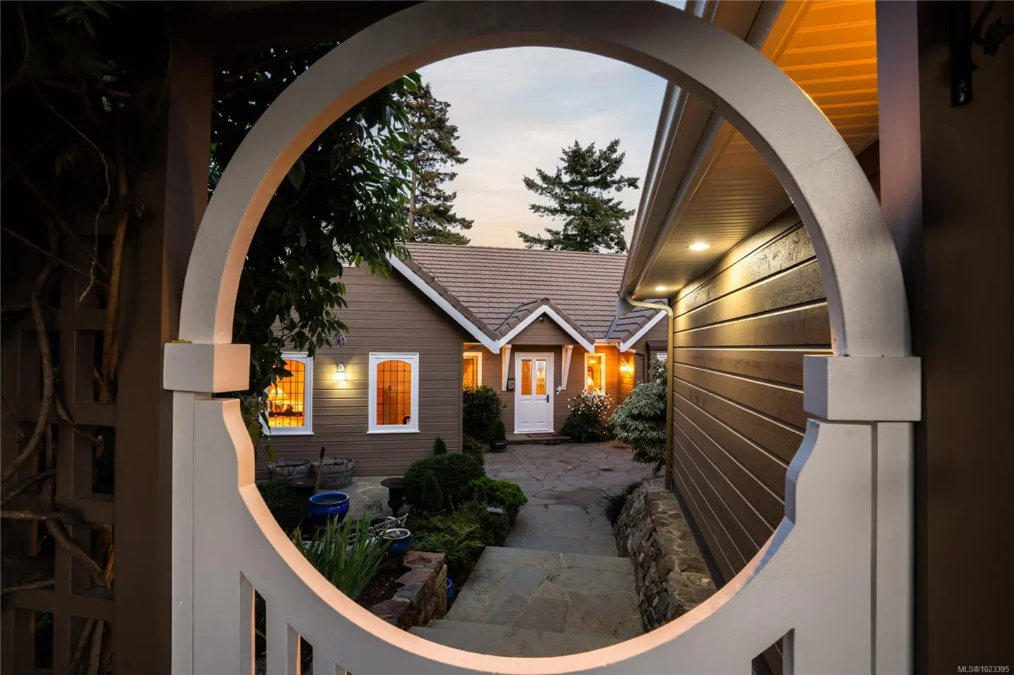 View of a brown house with a white door through a white circular archway. The windows glow with warm light.