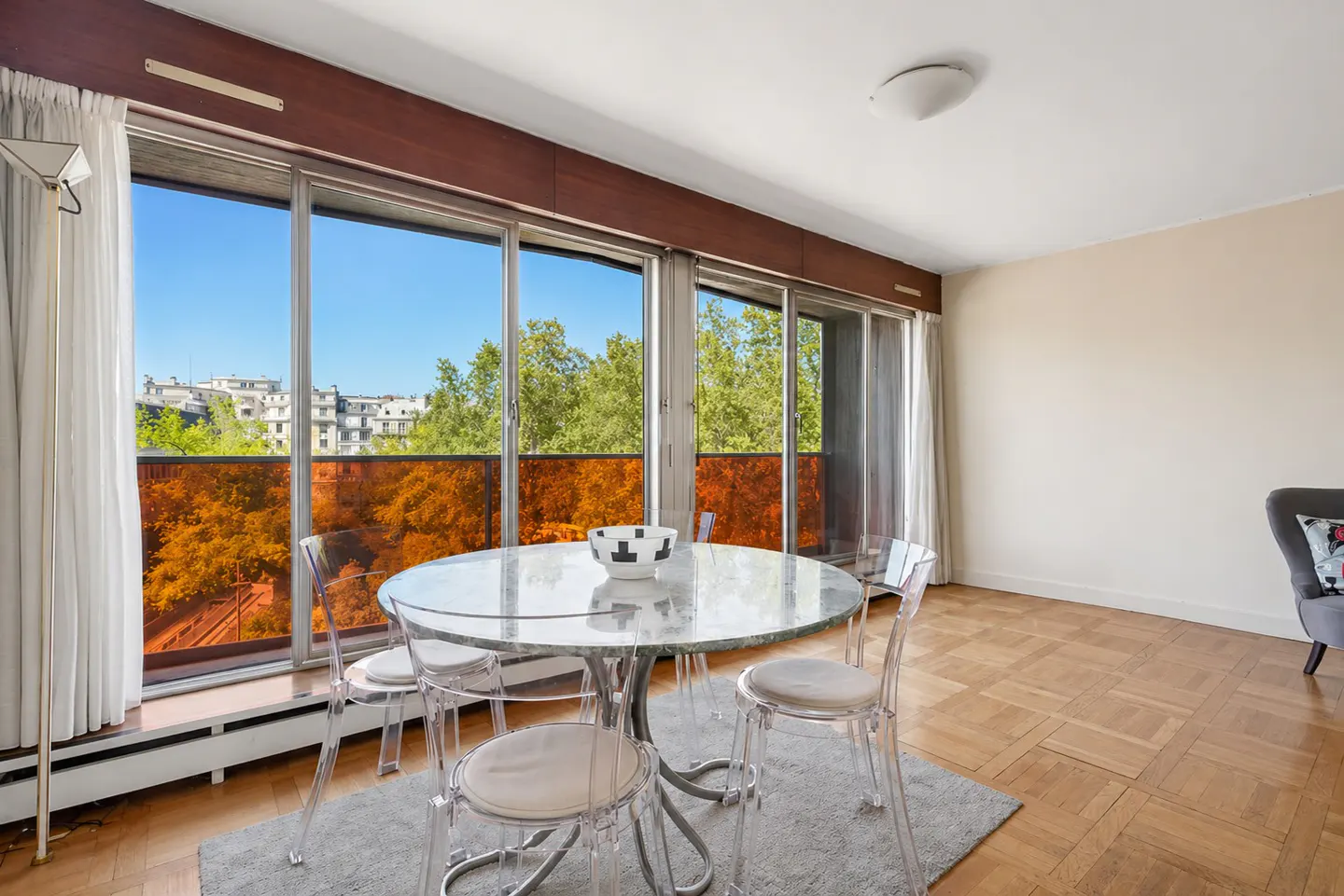 Bright dining room with parquet floors, a round marble table, and clear chairs. Large windows offer a view of trees and buildings.