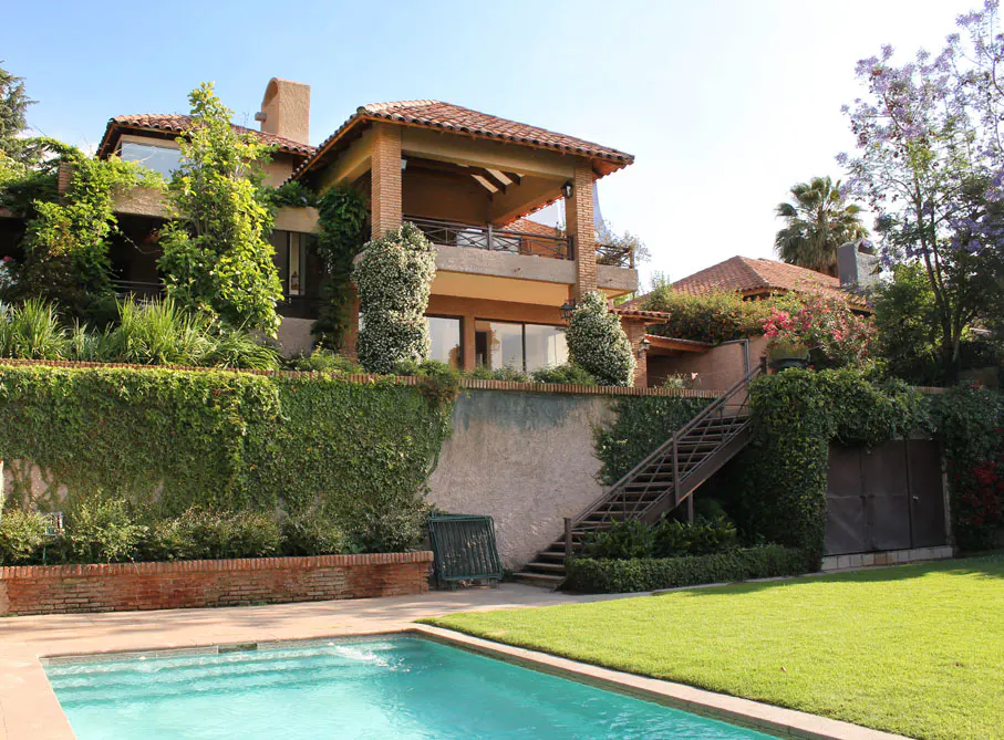 Two-story house with a red tile roof, a balcony, and a pool in the foreground. The house is surrounded by lush greenery.