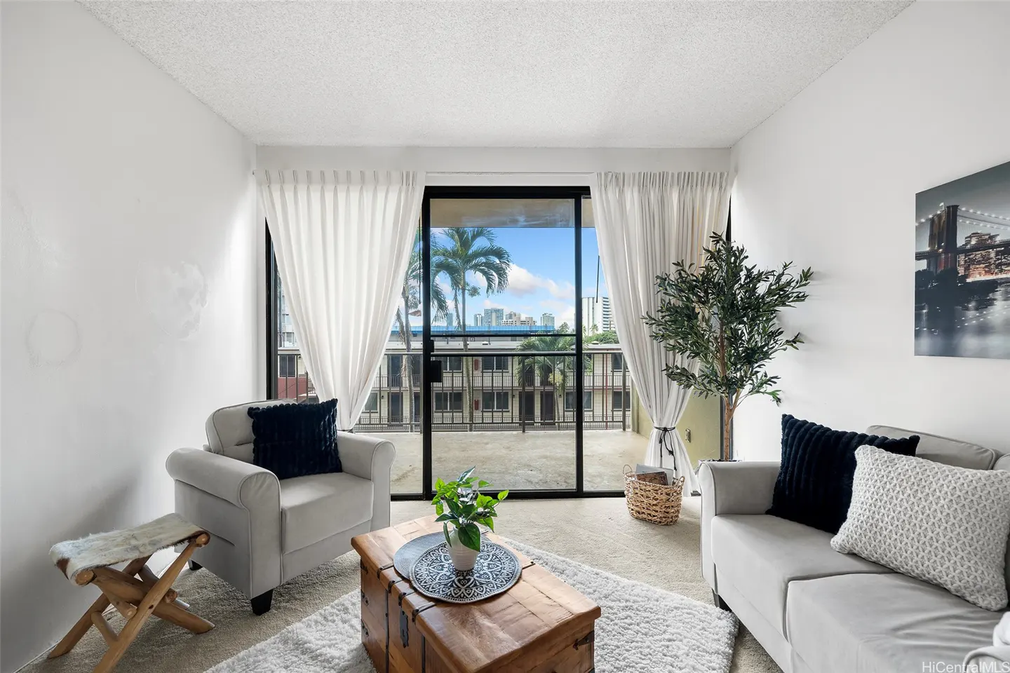 Living room with white walls, a gray sofa and armchair, a wooden chest as a coffee table, and a balcony with a view of palm trees and city buildings.